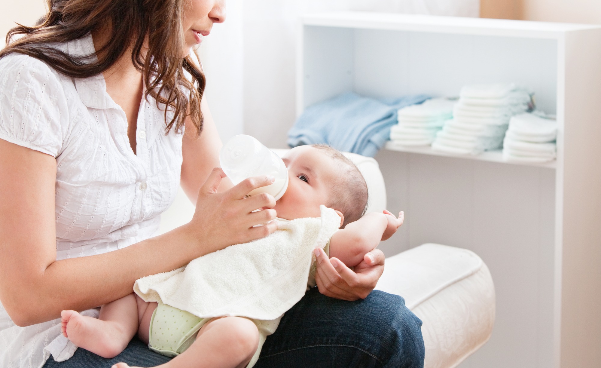 Close-up Of A Proud Mother Bottle-feeding Her Baby Sitting In The Sofa